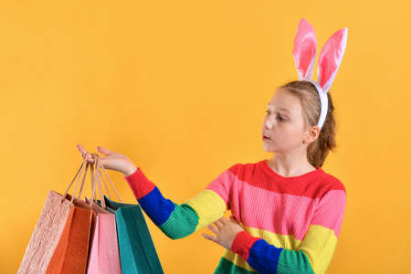 Girl with bunny ears and colorful clothes holds a shopping bag in her hand on a yellow background,...