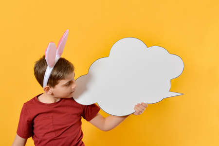 A boy with Easter ears holds in his hand a blank poster in the form of a cloud and looks at it. Advertising, text, advertisement, sale or purchase.の写真素材