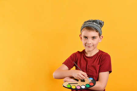 The boy holds a palette with paints and a brush in his hands, a child artist in a hat on a yellow background.の写真素材