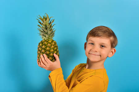 Pleasant and cute boy in a yellow T-shirt holds a pineapple in his hand, on a blue background.の写真素材