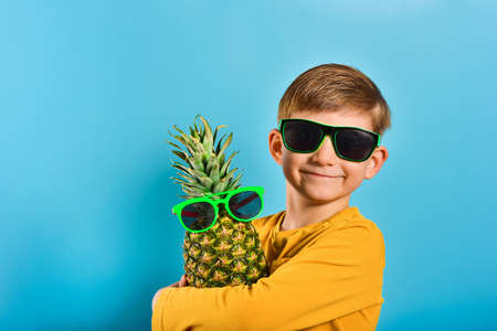 A nice and cute boy in a yellow T-shirt and sunglasses hugs a pineapple in glasses with his hands.