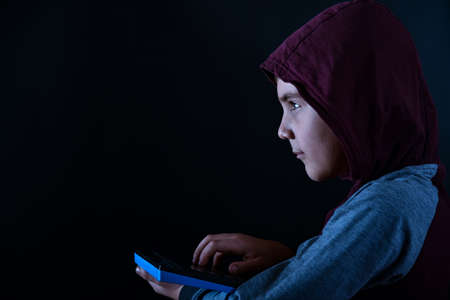 The boy in the hood, a young hacker, looks at the monitor, holding a keyboard in his hand.