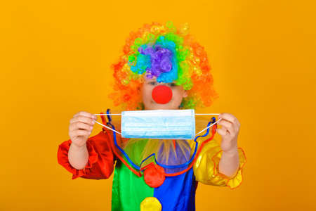 A sad and sad baby clown in a protective medical mask from a coronavirus holds a colored wig in one hand and the clowns nose in the other.の写真素材