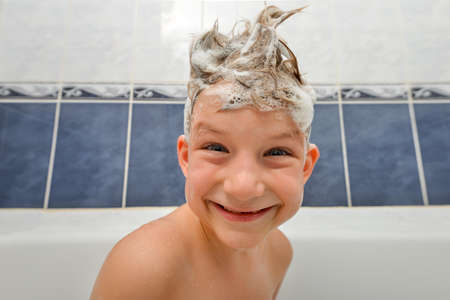 A child with a soapy head from shampoo sits in the bathroom in the shower room and smiles.