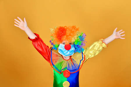 Joyful baby clown in a protective medical mask from a coronavirus on a yellow background holds his...
