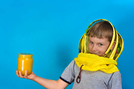 A boy in a protective mask from bees holds in his hand a jar of honey on a blue background.
