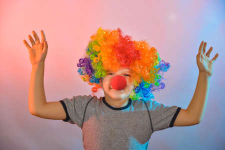 Joyful boy in a clown wig and with a red nose holds his hands to the sides.の写真素材