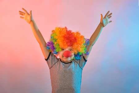 Joyful boy in a clown wig and with a red nose holds his hands to the sides.の写真素材