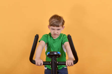 Portrait of a child on an exercise bike, the boy is engaged in cardio training and looking at the camera, on a yellow background.の写真素材
