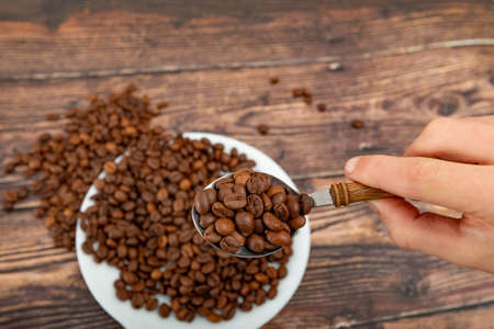 A man holds roasted coffee grains in a tablespoon on a background of a white plate and dark woodの写真素材