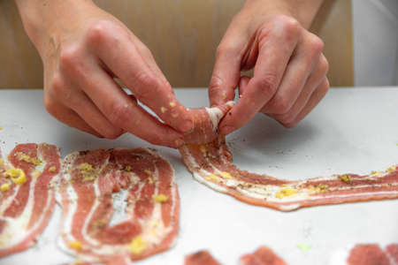 Woman hostess rolls thinly sliced bacon into roll for further cooking and frying in a pan. Cooking junk and fatty foodsの写真素材