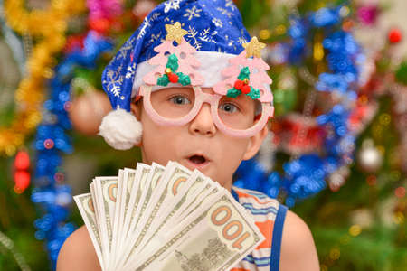 A boy in a blue Santa Claus hat holds out his hand with dollars, against the background of a Christmas tree. New year investment concept. Feast of Christmas.の写真素材