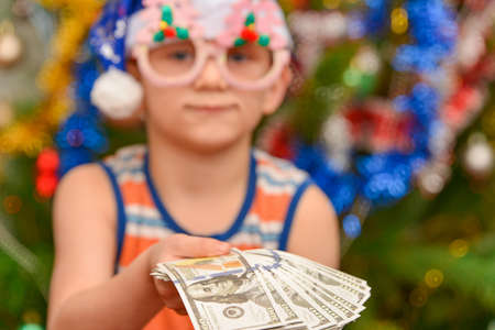 A boy in a blue Santa Claus hat holds out his hand with dollars, against the background of a Christmas tree. New year investment concept. Feast of Christmas.の写真素材