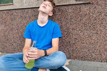 Drunk teenager with alcohol in his hands is sleeping sitting on the street and begging.の写真素材