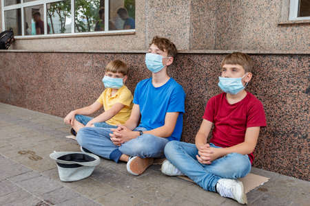 Three children wearing protective masks sit outside asking for financial assistance during the covid-19 coronavirus pandemic crisisの写真素材