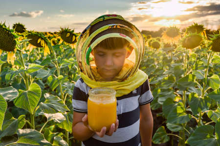 A boy wearing a beekeeper mask holds a jar of honey among a sunflower field and looks into it...
