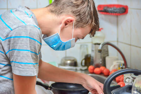 A surprised boy in a protective mask is engaged in washing dishes and preparing food in the kitchen at home, quarantined during the covid-19 pandemic.の写真素材