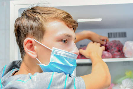 A boy wearing a protective mask takes food out of the refrigerator, quarantined during the covid-19 pandemic.の写真素材