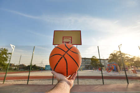 The hand of a male athlete holds a basketball ball on the background of a shield with a ring. Sports activities on the playgroundの写真素材