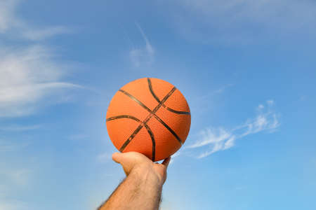 The hand of a male athlete holds a basketball ball against a blue cloudy sky. Sports activities on the playgroundの写真素材