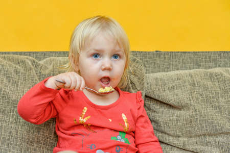 A little hungry girl eats porridge from a plate while sitting on the couch and watches children TV programs on TV.の写真素材