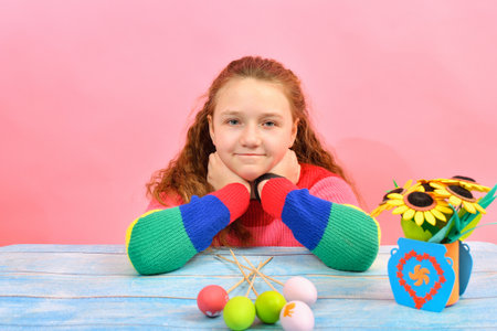 A joyful little girl sits at the Easter table with colored eggs on a pink background.