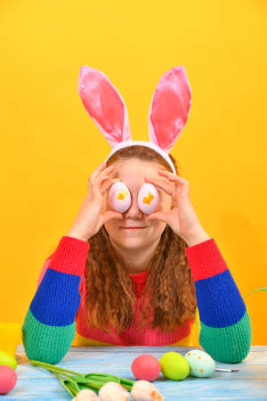 A girl with pink bunny ears holds Easter eggs on her face near her eyes.