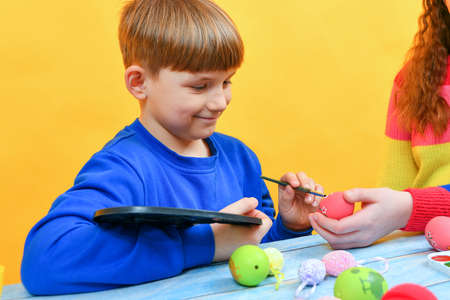 A boy paints Easter eggs with a paintbrush with the help of his older sister.の写真素材