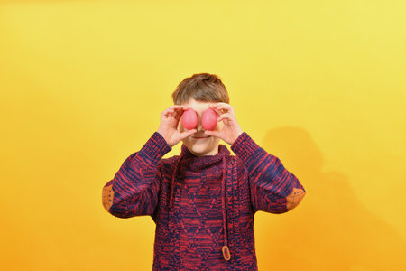 A boy in a red sweater holds Easter eggs in front of him on a yellow background.の写真素材