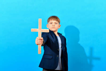 A boy in a suit holds a Catholic cross in front of him against a blue background.の写真素材