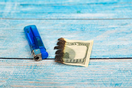 A burnt one hundred dollar bill and a blue lighter lie on a wooden texture.の写真素材