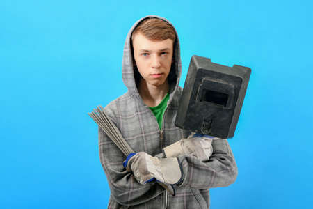 Young welder with electrodes and welding shield on a blue background.