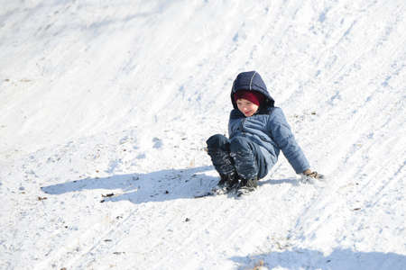 The boy sits on his haunches and descends on his feet on a slippery snow slide.