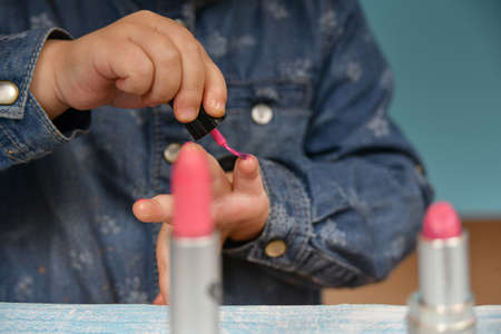 A little girl clumsily paints her nails with red varnish. Lipstick and other cosmetics are on the table.の写真素材