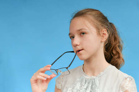 A young girl holds glasses in her hand and looks thoughtfully to the side. Portrait of a girl on a blue background.の写真素材