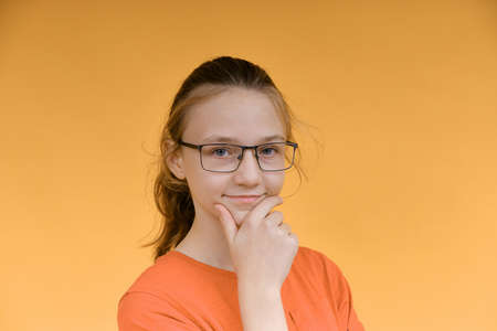A young girl in glasses holds her face with her hand and looks thoughtfully into the camera. Portrait of a girl on a yellow background.の写真素材