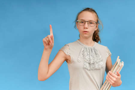 A girl in glasses with textbooks in her hand shows the index finger to the top.