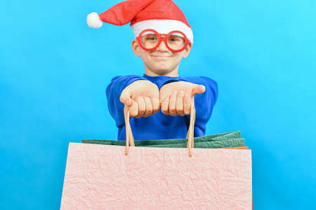 A joyful boy in funny glasses holds gift paper colored shopping bags in his hands. Shopping and...