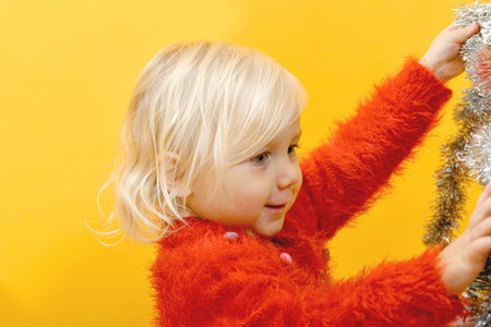 A little girl in a red sweater decorates the tree with festive toys and glittering tinsel.