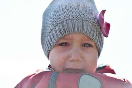 The child is crying in the street. Little girl in a hat and a red jacket with tears in her eyes.