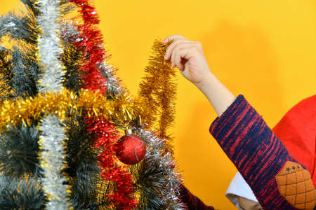 A boy wearing a red Santa Claus hat decorates a Christmas tree with festive decorations and toys.