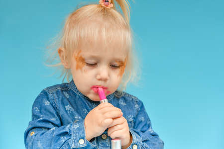 The little girl took the makeup from her mother and paints her lips with red lipstick.