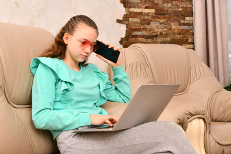 A young girl with a laptop sits in the office on a leather sofa and holds a phone in her hand.