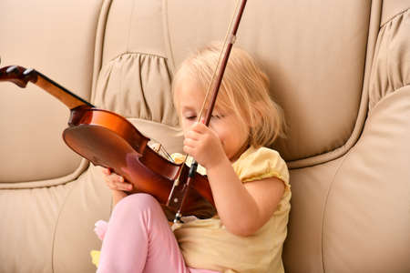 A little girl sits on a sofa with a violin and does not know what to do with it.の写真素材