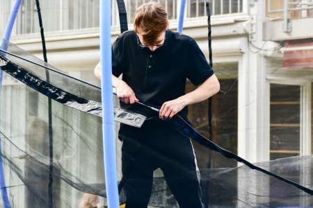 The guy collects a new trampoline at home and pulls on a protective net.