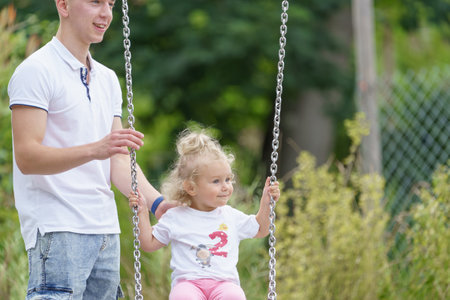 A young father pushes his little daughter on a swing in the park.の写真素材