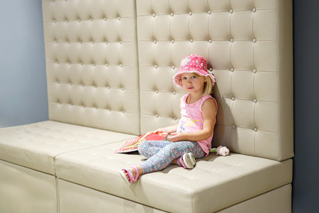 Little girl with a fashion magazine on a light leather sofa.