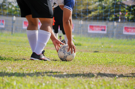 A dynamic, low-angle shot shows a football player, dressed in black shorts and white socks, placing an old, worn soccer ball on the green grass of a field. His hands are on the balの写真素材