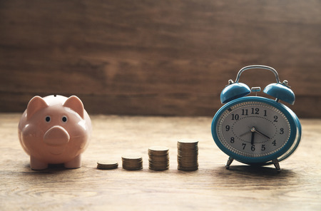 Piggy bank, stack of coins and alarm clock on wooden table. Time to invest your savings