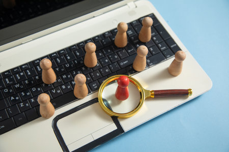 Magnifying glass, wooden human figures on laptop keyboard. Candidate selection